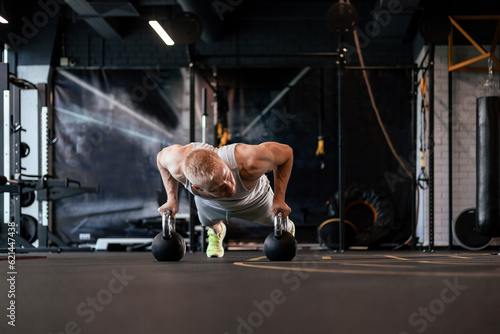 Fit and muscular man doing horizontal push-ups on dumbbells in gym.