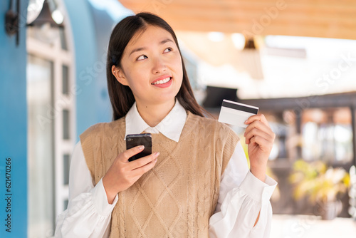 Young Chinese woman at outdoors buying with the mobile with a credit card while thinking photo