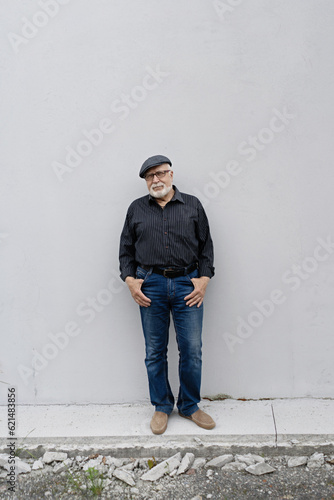 An elderly man in glasses and a cap stands on the background of a light wall, hands in his pockets, vertical format