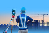 Surveyor with back to camera. Man near geodetic instrument. Worker looks at high-rise building under construction. Surveyor man with theodolite. Geodetic apparatus on tripod. Geodetic business