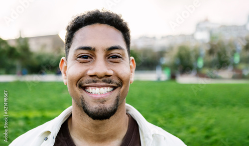 Outside smiling portrait of young latino hispanic gen z man looking at camera standing at city park. Front headshot of joyful student guy laughing outdoors.