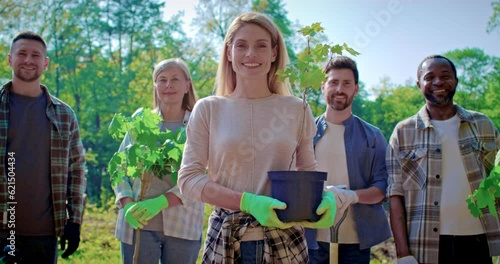 Portrait of volunteer woman holding plant in forest on background of volunteers. Young woman holds in hands plant sprout. Voluntary free work assistance help, charity grace concept. photo