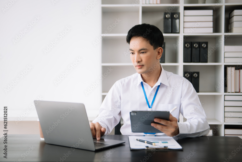Young Asian business man using laptop and tablet while sitting at her working place. Concentrated at work. Young Asian business man using laptop and tablet while sitting at her working place. Concentrated at work.