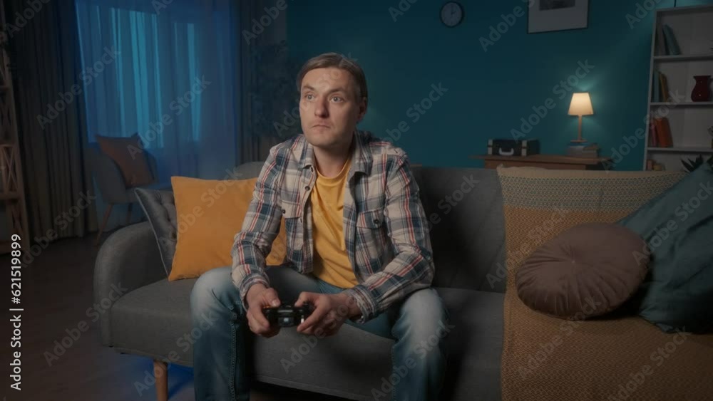 A man plays a consular game with a game controller while sitting on the couch at home in the evening. The man is nervous, loses, gets angry, throws away the joystick. A man plays a consular game with a game controller while sitting on the couch at home in the evening. The man is nervous, loses, gets angry, throws away the joystick.