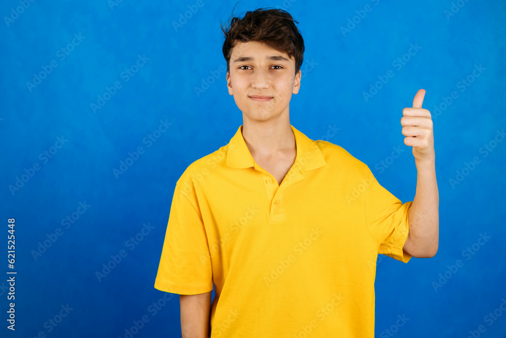 Close-up portrait of a teenager in a yellow T-shirt on a blue background, smiling, looking at the camera, pointing a finger up. Copy space Close-up portrait of a teenager in a yellow T-shirt on a blue background, smiling, looking at the camera, pointing a finger up. Copy space
