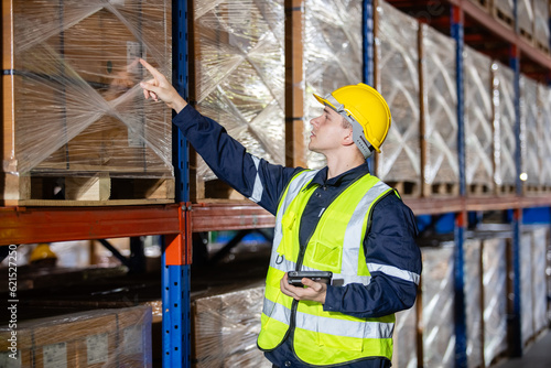 Male professional worker wearing safety uniform and hard hat using digital tablet inspect product on shelves in warehouse. Man worker check stock inspecting in storage logistic factory.