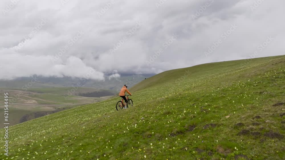 A male cyclist rider in a protective helmet on a mountain bike rides on green hills high in the mountains against the backdrop of clouds and mountains. aerial view A male cyclist rider in a protective helmet on a mountain bike rides on green hills high in the mountains against the backdrop of clouds and mountains. aerial view