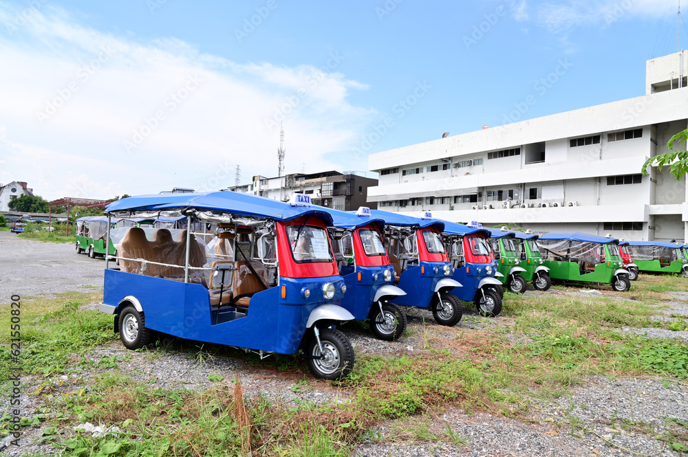 BANGKOK, THAILAND - JULY 10, 2023: Several Electric tuk tuks taxi stopped in the meadow with buildings background. Concept to save fuel, maintain and reduce air pollution protect the environment.