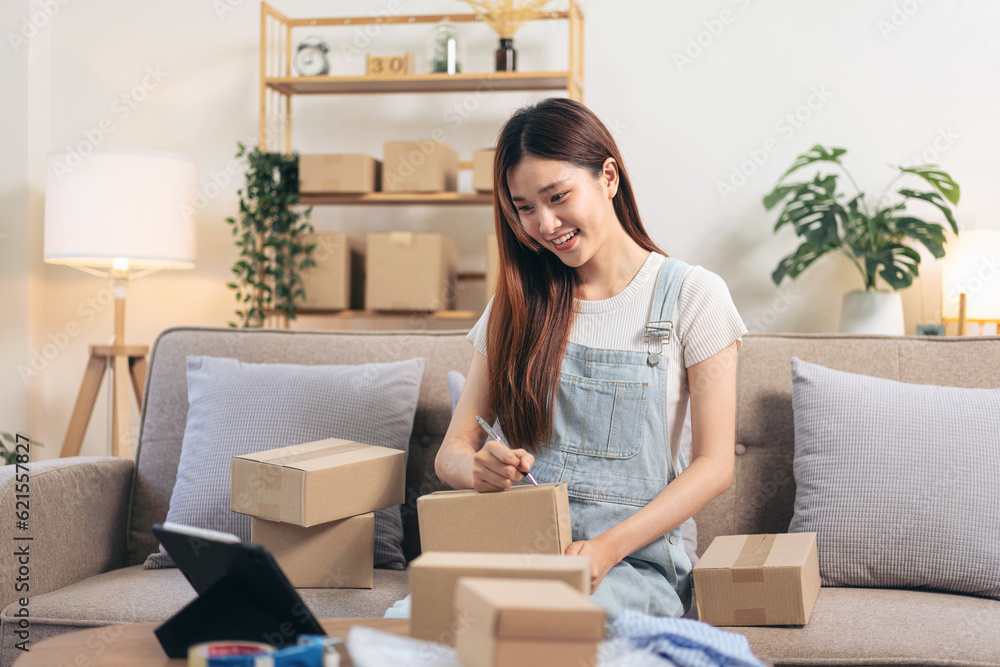 Young entrepreneur is using tablet to checking online orders and writing customer address on cardboard boxes Young entrepreneur is using tablet to checking online orders and writing customer address on cardboard boxes