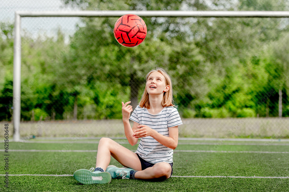 Foto de Pretty child girl sitting on grass and throwing football ball up. Cute female kid at