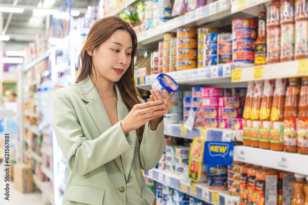 Asian young woman, female in grocery, supermarket shopping food store hand holding tuna can, take out of shelf, read expired date information and ingredients on label, buying healthy products in mall