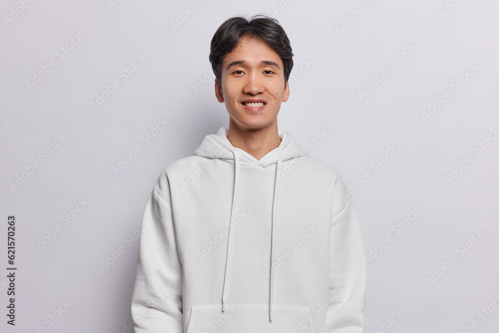 Portrait of handsome Asian man with dark hair dressed in casual sweatshirt smiles pleasantly looks directly at camera expresses positive emotions isolated over white background. Monochrome shot Portrait of handsome Asian man with dark hair dressed in casual sweatshirt smiles pleasantly looks directly at camera expresses positive emotions isolated over white background. Monochrome shot