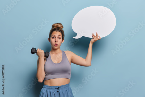 Horizontal shot of sporty healthy woman dressed in cropped top and shorts lifts dumbbell holds blank speech bubble for your advertising content isolated over blue background. Fitness nutrition concept