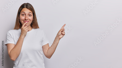 Young European woman wearing white tshirt directs attention to upper right corner where good offer awaits encourage viewers to click on link dressed in basic tshirt isolated over white background