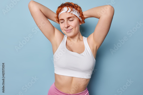Horizontal shot of redhead woman keeps hands behind head stands with closed eyes rests after workout smiles gently isolated over blue background. Sporty lifestyle and regular training concept