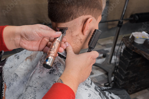 The hands of a man of an experienced hairdresser hold a brush for hair removal and cut the back of the head, make a haircut for a young guy client at work