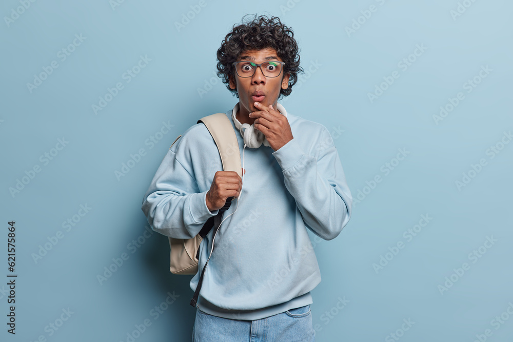 Indoor shot of curly haired Hindu man stares through transparent eyeglasses keeps lips folded dressed in casual pullover and jeans carries rucksack isolated over blue background. People and lifestyle Indoor shot of curly haired Hindu man stares through transparent eyeglasses keeps lips folded dressed in casual pullover and jeans carries rucksack isolated over blue background. People and lifestyle