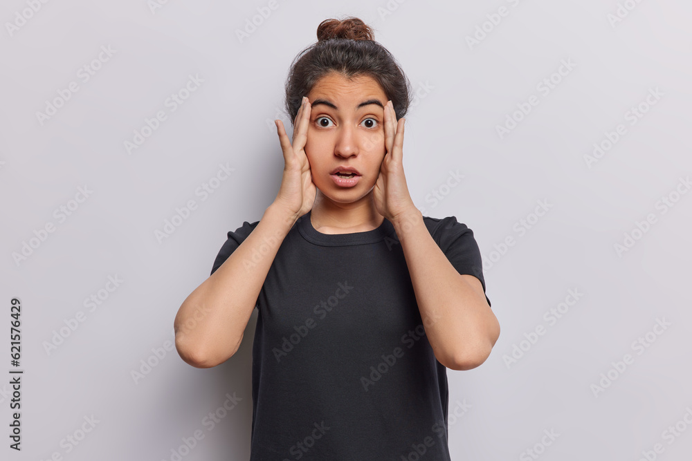 Studio shot of stunned dark haired woman looks with terror keeps hands on head notices something horrible dressed in casual black t shirt isolated over white background. Human reactions concept Studio shot of stunned dark haired woman looks with terror keeps hands on head notices something horrible dressed in casual black t shirt isolated over white background. Human reactions concept