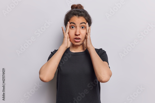 Studio shot of stunned dark haired woman looks with terror keeps hands on head notices something horrible dressed in casual black t shirt isolated over white background. Human reactions concept