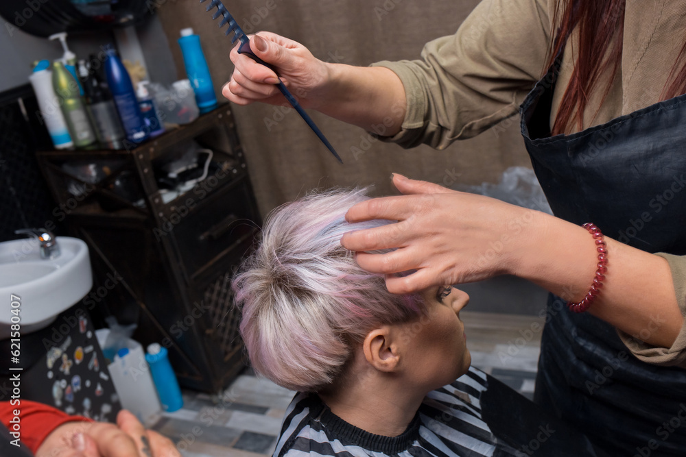 The hands of a girl of an experienced salon worker with a comb correct the hairstyle of a client to an adult woman with stylish, colored hair coloring