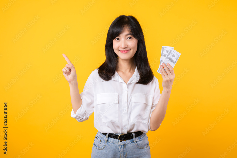 Young happy woman of Asian ethnicity wear white shirt and denim jean holding cash money in dollar and pointing finger to free copy space against yellow background. Investment and financial concept. Young happy woman of Asian ethnicity wear white shirt and denim jean holding cash money in dollar and pointing finger to free copy space against yellow background. Investment and financial concept.