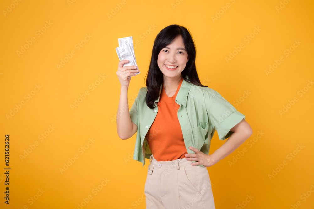 Asian woman 30s holding currency dollar while confidently placing hand on hip isolated on yellow background, embodies financial empowerment and style. financial stability and personal success concept. Asian woman 30s holding currency dollar while confidently placing hand on hip isolated on yellow background, embodies financial empowerment and style. financial stability and personal success concept.