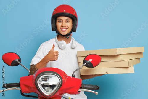 Happy asian boy in red helmet sits on scooter on blue background in studio, holds pizza boxes and smiles widely, fast delivery concept, copy space
