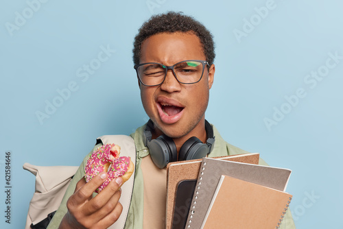 Fotografia Horizontal shot of tired university student yawns and keeps mouth opened wants to sleep after sleepless night poses with notepads and glazed sweet doughnut isolated over blue studio background