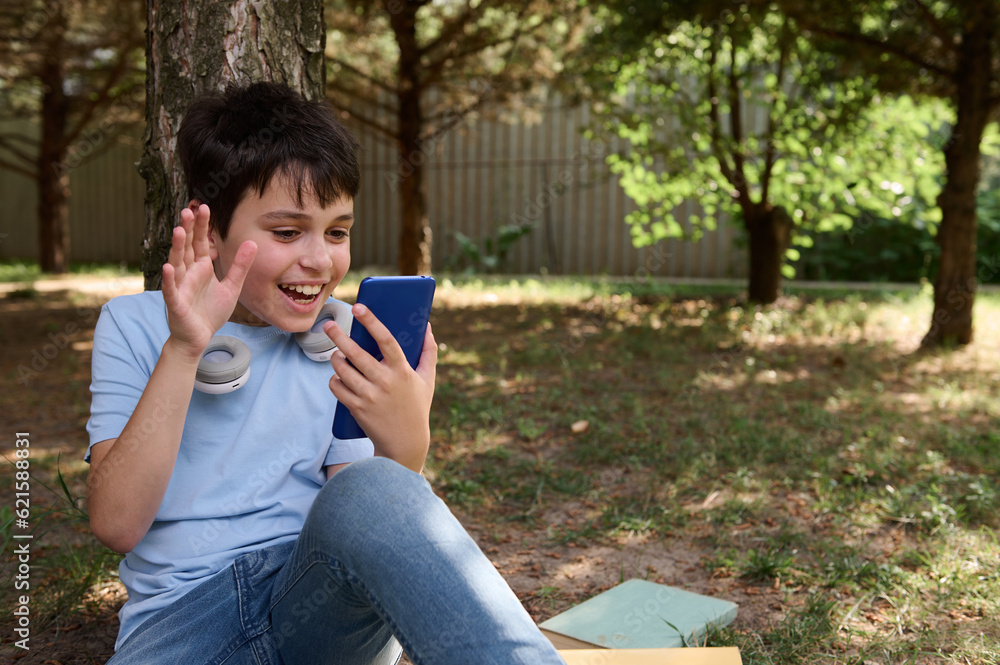 Cheerful teen boy waving hello with hand, having online conference by video link on his smartphone, sitting under a tree Cheerful teen boy waving hello with hand, having online conference by video link on his smartphone, sitting under a tree