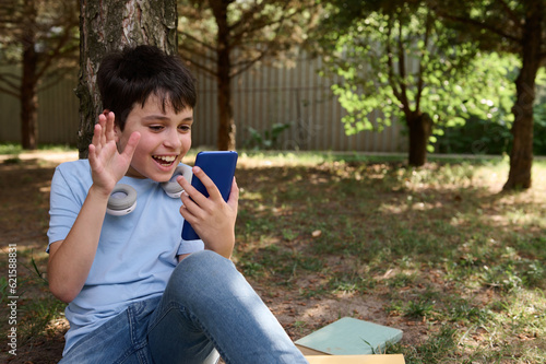 Cheerful teen boy waving hello with hand, having online conference by video link on his smartphone, sitting under a tree