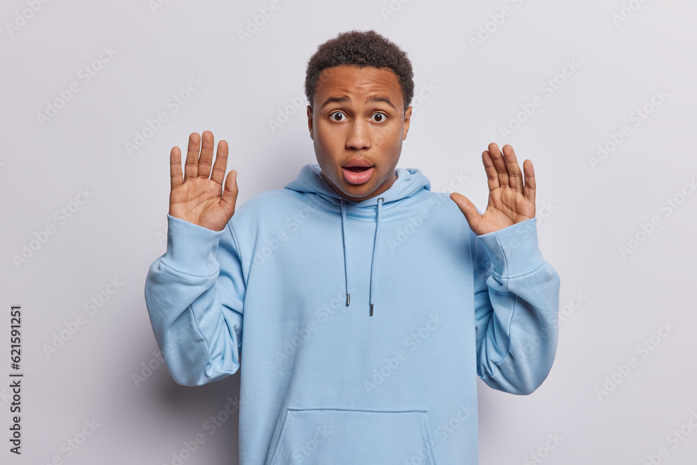 Photo of dark skinned curly haired man keeps palms raised up feels scared dressed in casual blue sweatshirt cannot believe in something shocking isolated over white background. Human reactions concept Photo of dark skinned curly haired man keeps palms raised up feels scared dressed in casual blue sweatshirt cannot believe in something shocking isolated over white background. Human reactions concept
