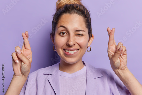 Photo Positive European woman winks eye exudes hope and optimism as she crosses her fingers winks mischievously and flashes broad smile dressed in shirt isolated over purple background