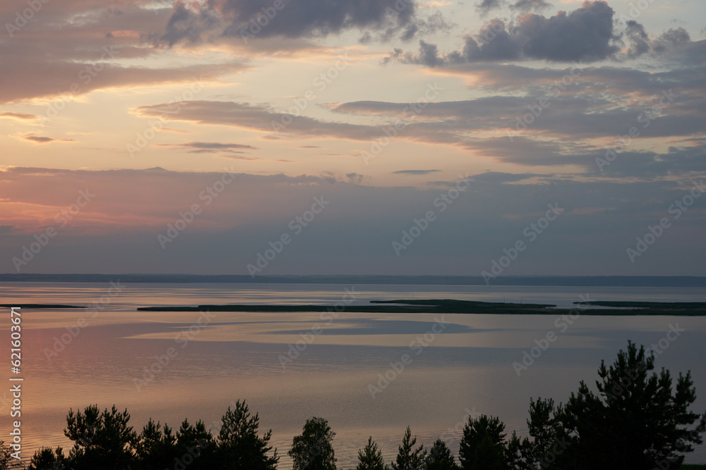 A wide plain river at sunset. The sky and the water surface of the river are colored golden pink. There are ripples on the water. Silence and tranquility. Copy space.