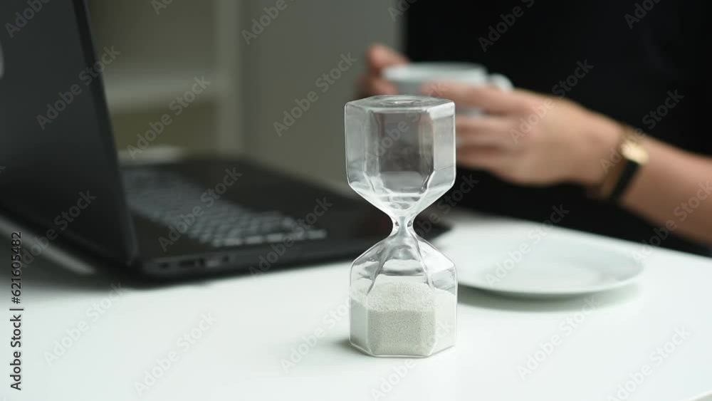 woman holds a cup in her hands, sitting in front of a laptop. There is an hourglass in the foreground, the background is blurred. Study, business, work, communication via PC woman holds a cup in her hands, sitting in front of a laptop. There is an hourglass in the foreground, the background is blurred. Study, business, work, communication via PC