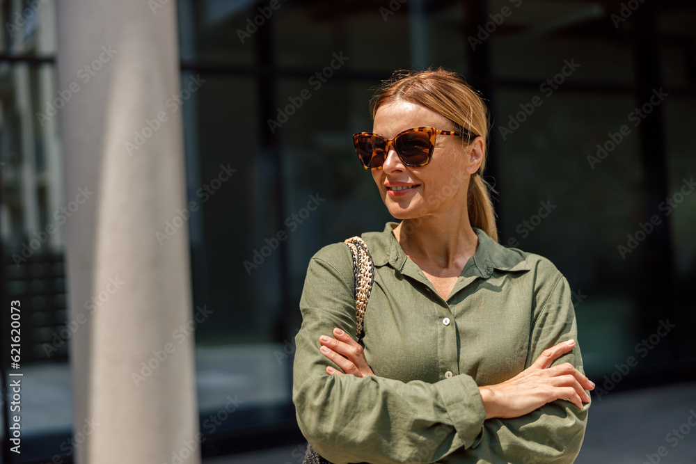 Fototapeta premium Businesswoman with crossed hands is standing on office building background and looking at side
