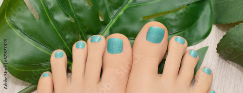 Female feet with blue glitter nail design. Blue nail polish pedicure and greel leaves on background.