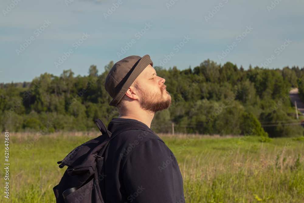 A young bearded Caucasian male traveler with a backpack in a panama stands in a field with his eyes closed in the sunlight against the forest on a warm summer day. The concept of a happy life