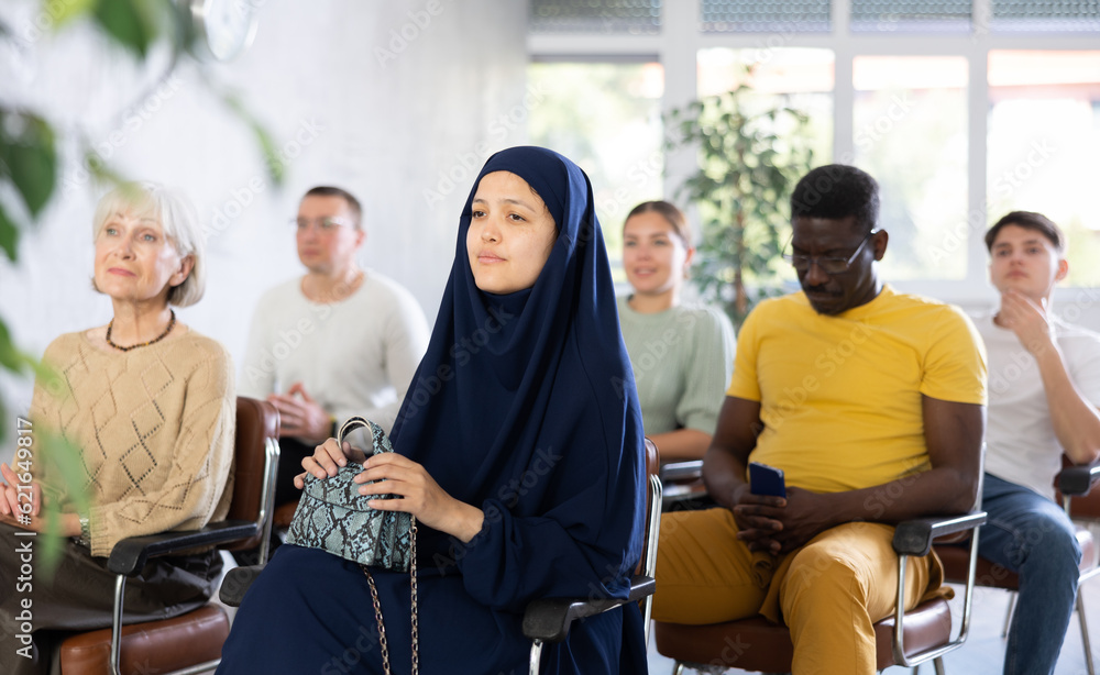 Naklejka premium Focused young asian girl dressed in traditional muslim abaya and chador sitting in lecture hall with group of men and women of different ages