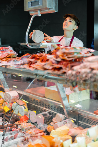 Young man in apron working in butchery, standing at counter, using scale to weigh sausage.