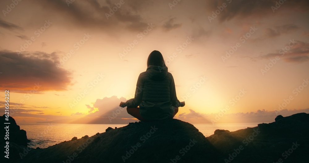 Silhouette of relaxed woman against sunset sky on rocky beach at sunset. Girl meditates in nature and sun rays illuminate her.