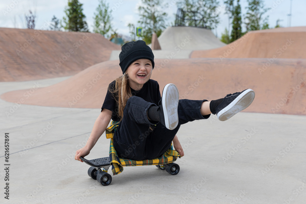 Pretty happy little girl sits on a skateboard on the street in the summer. Children's sports summer. Hobbies at home Pretty happy little girl sits on a skateboard on the street in the summer. Children's sports summer. Hobbies at home