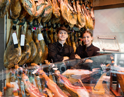 Portrait of positive young man and woman jamoneria workers standing at counter with arms crossed, looking at camera and smiling.
