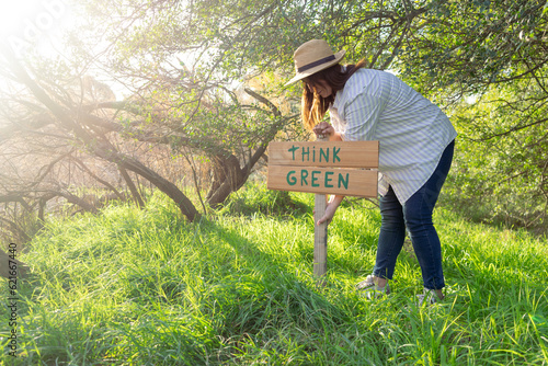 young woman with a hat in an environment of trees and sun with a wooden sign that says think green to protect the natural environment and take care of the environment photo