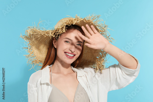 Beautiful young woman in straw hat shading herself with hand from sunlight on light blue background photo