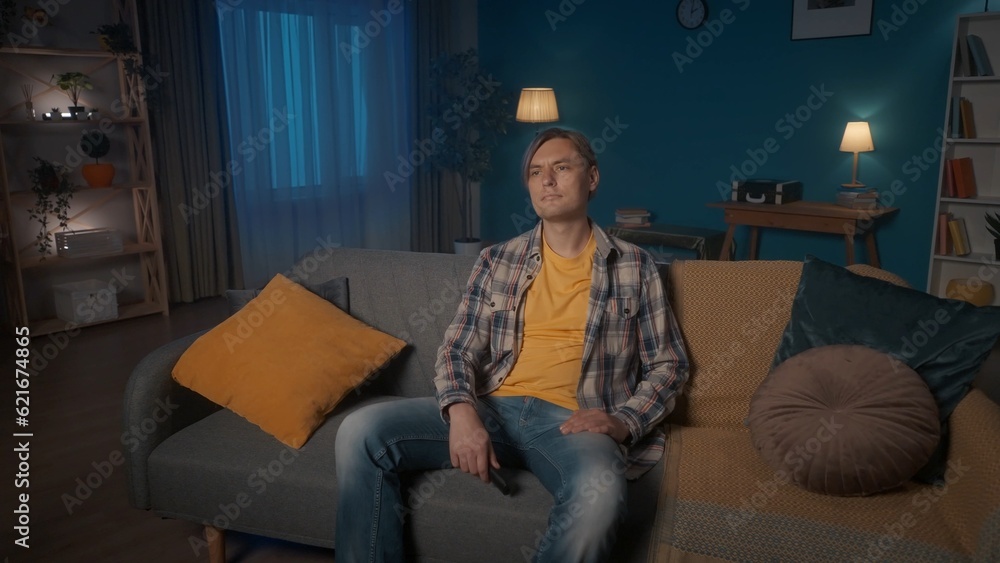 Fototapeta premium A young man sits on the sofa in the evening in the room and watches TV. A man is sitting with a TV remote control.