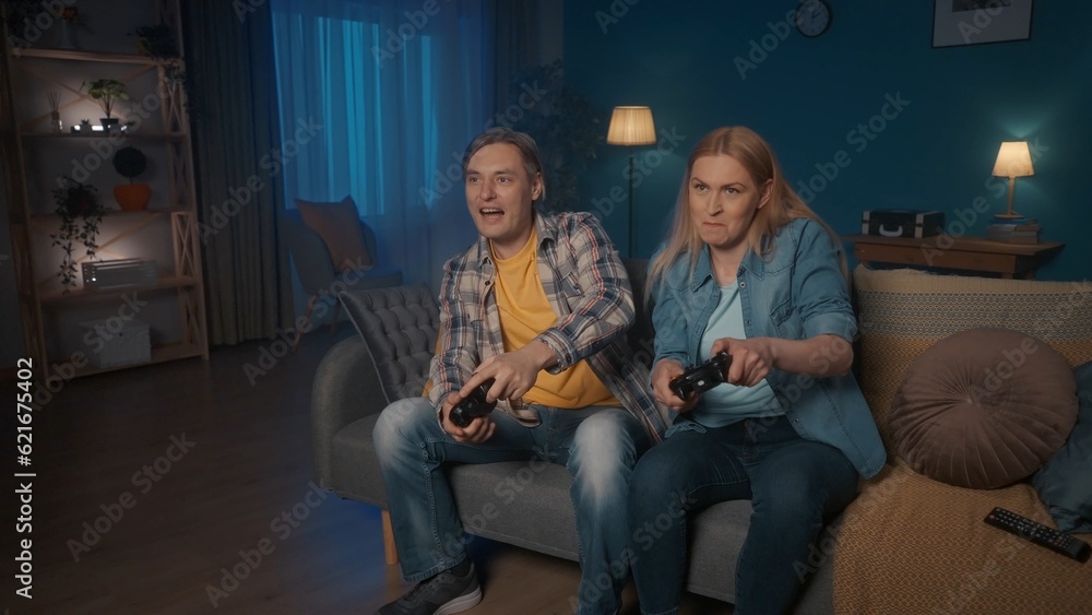 A young couple is playing a video game console with game controllers while sitting on the couch at home in the evening. Man and woman spend time together enjoying the game. Who will win. Game concept