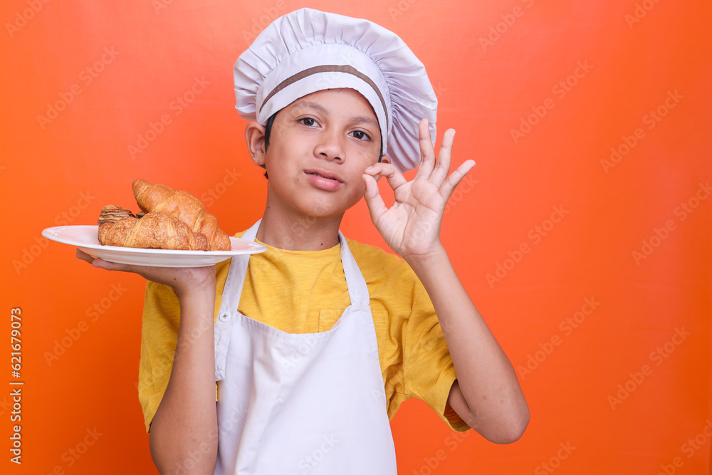 Cute adorable liitle boy in chef uniform and apron holds plate with croissant bread showing delicious sign Cute adorable liitle boy in chef uniform and apron holds plate with croissant bread showing delicious sign