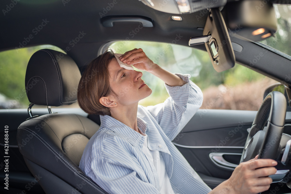 Obraz premium Woman drives car with broken air conditioning in hot summer weather wipes sweat paper napkin. Middle aged female suffering from heat stuffiness. Tired exhausted overheated stressed worried driver.