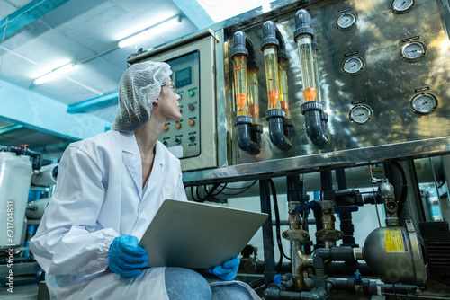 Female worker using laptop checking quality drinking water management system before process of filling water into bottles to bring out to consumers. Water production line of factory.