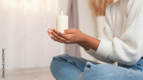 Wish dream. Candle praying. Spiritual worship. Unrecognizable woman holding burning flame wax light in hands sitting lotus pose at home. photo